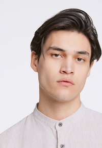 Camisa de botón color beige claro con cuello tipo banda, hecha de una tela texturizada. Cuenta con dos botones en la parte delantera y un diseño ligeramente relajado.