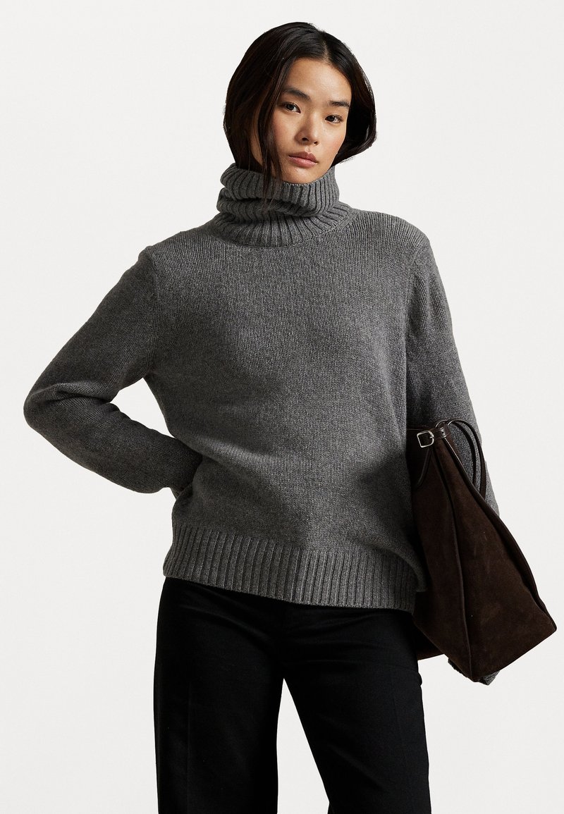 Pelēks turtleneck džemperis, kas izgatavots no bieza adījuma auduma, ar ribotu apkakli un apakšu, saskaņots ar melniem biksēm un tumši brūnu somu.