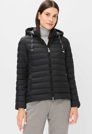 Woman wearing a black quilted hooded jacket over a gray turtleneck and light gray pants, standing with relaxed arms.