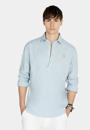 Młody mężczyzna w jasno niebieskiej koszulce polo z długim rękawem z podwiniętymi rękawami i białych spodniach, stoi z rękami w kieszeniach.