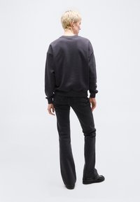 Άτομο με κοντά ξανθά μαλλιά που φοράει σκούρο γκρι φούτερ, μαύρα καμπάνα τζιν και μαύρα παπούτσια, στέκεται γυρισμένος προς τα πίσω σε λευκό φόντο.