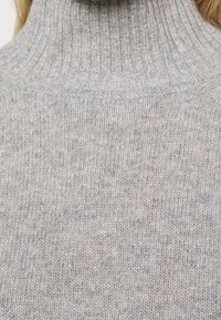 Lysegrå rullekravetrøje lavet af strikket stof. Har en høj krave og en blød, struktureret overflade med et subtilt mønster.