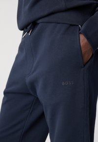 Tumsā zilas sporta bikses, izgatavotas no mīksta auduma, ar elastīgu jostu, regulējamu auklu, sānu kabatām un diskrētu "BOSS" logotipu.