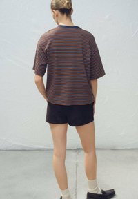 Pruhovaná košeľa s krátkym rukávom v hnedej, modrej a oranžovej farbe, skombinovaná s tmavými šortkami. Tkanina pôsobí mäkko a ležérne, s voľným strihom.