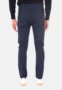 Uomo che indossa pantaloni skinny blu navy, maglietta nera a maniche lunghe e scarpe da ginnastica bianche, in piedi con la schiena rivolta verso uno sfondo neutro.