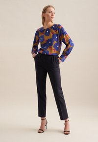 Blusa a maniche lunghe con motivo floreale blu, giallo e rosa, abbinata a pantaloni neri su misura e tacchi alti neri con cinturini. Texture liscia.