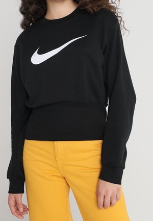 Person trägt einen schwarzen Nike Pullover mit langen Ärmeln und weißem Swoosh-Logo sowie hoch taillierte gelbe Hosen vor einem einfarbigen Hintergrund.