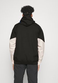 Personne aux cheveux bouclés portant un sweat à capuche noir avec des manches beige et un pantalon foncé, debout de dos sur un fond uni.