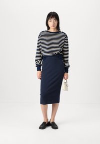 Pull rayé marine avec des boutons dorés, associé à une jupe marine ajustée. Porté avec des chaussures noires à enfiler et un petit sac à main blanc.