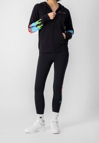 Felpa nera con zip e maniche tie-dye colorate, abbinata a leggings neri con una grafica colorata e scarpe da ginnastica bianche.