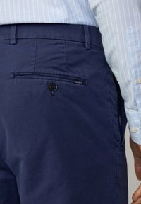 Chino blu navy con una texture liscia, dotati di una tasca posteriore con bottone e un piccolo etichetta del marchio con la scritta "HACKETT". Indossato con una camicia a righe azzurra.