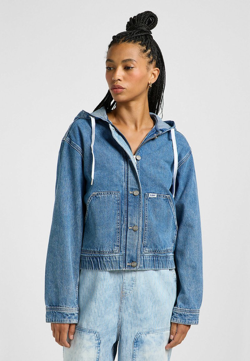 Lyhyt denim-takki keskisinisessä sävyssä, jossa on kiristysnauha huppu, etuvaatetus ja kaksi suurta läpällistä taskua. Pehmeä pinta, rento muotoilu.