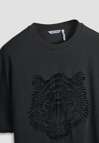 Camiseta de algodón negra con un diseño texturizado de la cara de un león en tela negra. Presenta un cuello redondo clásico y detalles de costura sutiles.