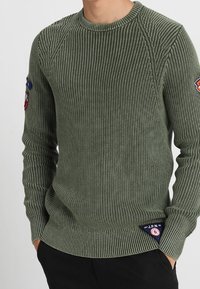 Man in een olijfgroene geribbelde gebreide trui met patches op de mouwen en een kleine driehoekige patch bij de zoom, met handen in de zakken van zijn zwarte broek.