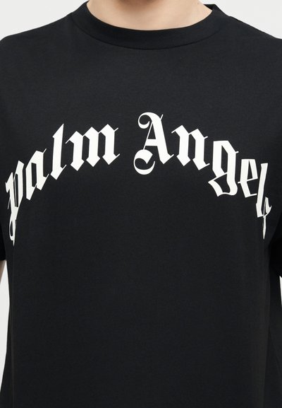 Μαύρο μπλουζάκι crewneck με τη φράση "Palm Angels" σε μεγάλο λευκό γοτθικό γράμμα στο στήθος, φορεμένο από άτομο που εμφανίζεται από το λαιμό έως το μέσο του κορμού.