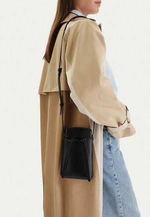 Žena nosící béžový trenčkot a světle modré džíny, s malou černou crossbody taškou s tenkými raménky.
