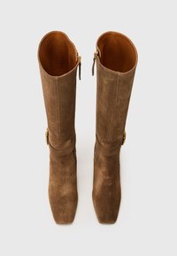 Botas de camurça castanhas até ao joelho com biqueira quadrada, pormenores em metal dourado e fecho de correr na parte de trás. Interior forrado em couro bege.