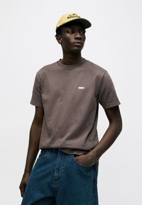 Νέος άνδρας φορώντας καφέ μπλουζάκι OBEY, μπλε τζιν και κίτρινο καπέλο μπέιζμπολ, στέκεται με το ένα χέρι στην τσέπη του μπροστά σε μια απλή φόντο.