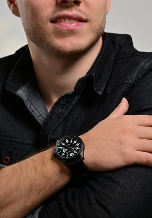 Schwarze analoge Uhr mit großen weißen Ziffern auf einem runden Zifferblatt, schwarzem Kunststoffgehäuse und strukturiertem Stoffarmband, getragen am Handgelenk über dunklem Stoff.