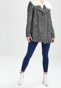 Un abrigo peacoat de tweed en blanco y negro con un cuello de piel sintética, combinado con unos vaqueros azules y botas de tobillo blancas con tachuelas doradas.