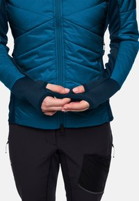 Teal gestepte jas met een volledige rits, duimopeningen en donkerblauwe accenten. Gecombineerd met zwarte broek met een getextureerde zak.