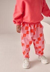 Jaskrawo różowe spodnie z pomarańczowymi kwadratowymi wzorami i elastycznym paskiem w talii, zestawione z różową bluzą i białymi trampkami.