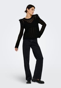 Kvinne iført svart blondetop med lange ermer og volanger på skuldrene, svarte vide jeans og svarte mokkasiner, poserer med hånden på hoften.