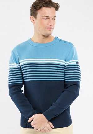 Homme portant un pull à manches longues à rayures horizontales bleu clair, blanc et marine, avec des boutons sur l'épaule, debout les mains jointes.