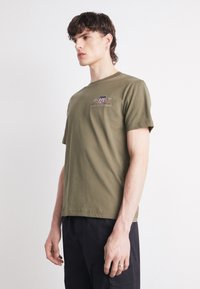 Camiseta de algodón verde oliva con mangas cortas, escote redondo, detalle del logo en el frente y corte holgado. Usada con pantalones negros.