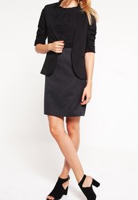 Blazer noir sur un top plissé noir, associé à une jupe noire texturée et des talons ouverts noirs. Design ajusté et cintré.