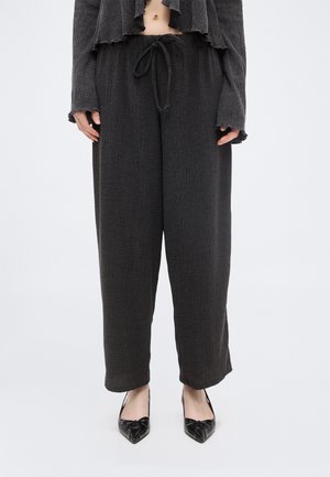 Személy laza, fekete, texturált nadrágot visel, melynek derékja húzózsinóros, és fekete hegyes orrú cipőt, egyszerű háttér előtt áll.