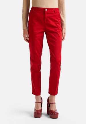 Woman wearing slim-fit red pants paired with beige sweater and red high-heeled Mary Jane shoes, standing against a plain background.