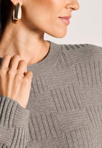 Frau trägt einen strukturierten grauen Pullover mit gerippten Quadratmustern und großen goldenen Tropfenohrringen, während sie den Kragen des Pullovers in der Nähe ihres Halses hält.