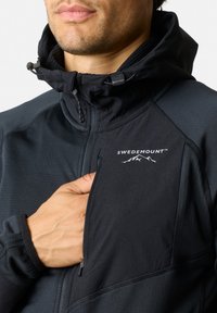 Zwart softshell jack met een hoge kraag, voorzien van een ritssluiting op de borstzak, elastische manchetten en een logo aan de bovenste linkerzijde.