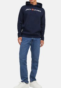 Marineblauer Hoodie mit weißem "JACK & JONES"-Schriftzug, einer Fronttasche und einer Kapuze mit Kordelzug, kombiniert mit blauen Jeans und weißen Sneakern.