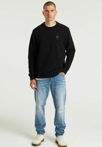 Schwarzes geripptes Sweatshirt mit Rundhalsausschnitt, kombiniert mit hellblauen verwaschenen Jeans und weißen Sneakers mit braunen Sohlen.