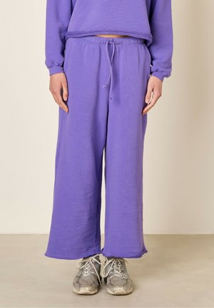 Lila Baumwoll-Sweatpants mit lockerer Passform, elastischem Bund und Kordelzug. Weicher Stoff mit rohem Saum-Detail. Kombiniert mit grauen Turnschuhen.
