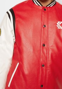 Röd läder varsityjacka med svarta och vita ärmar, tryckknappar, ribbad krage, vita detaljer och broderad logotyp.