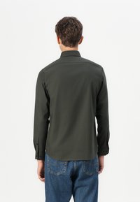 Mørkegrønn langermet skjorte med spiss krage, knappeledd og en glatt tekstur. Brukt med blå denim jeans.