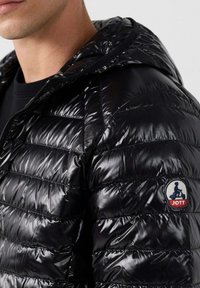 Giacca a vento nera con finitura lucida, dotata di sezioni trapuntate e cappuccio. Un badge circolare con logo blu e rosso è presente sulla manica.