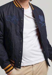Veste bombers en denim foncé avec des poignets côtelés rayés jaunes et noirs, arborant des broderies de dragon et de crâne sur la poitrine.
