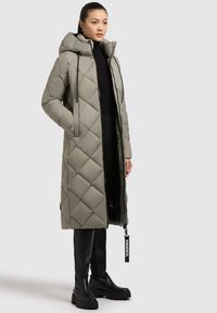 Cappotto lungo imbottito in grigio chiaro con cappuccio, chiusura a zip e cordini. Abbinato a stivali neri alti fino al ginocchio e a un top scuro.