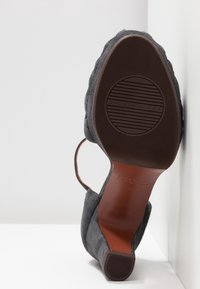 Semelle d'une chaussure à talon haut en daim gris avec une semelle en caoutchouc et une bride, inclinée contre un angle blanc entre un mur et le sol.