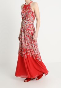 Robe longue rouge à motif floral avec un col noué autour du cou, présentant des motifs complexes et une jupe fluide. Accentuée par une texture et un tissu superposé.