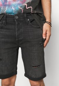 Pantalones cortos de mezclilla negros con agujeros deshilachados, puños doblados y diseño de cinco bolsillos. La tela tiene una textura ligeramente desgastada, mostrando signos de uso.