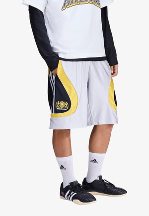 Hombre con pantalones cortos de baloncesto blancos y amarillos con paneles laterales negros, calcetines blancos de Adidas y zapatillas negras de Adidas, de pie con las manos en los bolsillos.