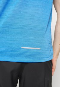 Kortærmet atletisk T-shirt i gradient blå stof, med struktureret mønster og en reflekterende pink detalje nær kanten. Sorte shorts er synlige.