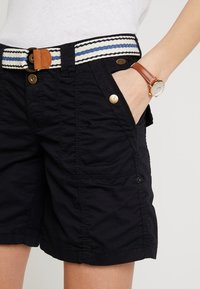 Personne portant un short noir avec des boutons dorés, une ceinture blanche et bleue rayée, et une montre en cuir marron avec un cadran blanc, main dans la poche.