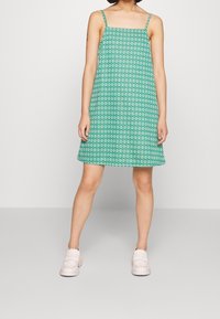 Zelené vzorované šaty bez rukávů s volným střihem, tenkými ramínky a geometrickým designem. Nosí se s světle růžovými pantoflemi.