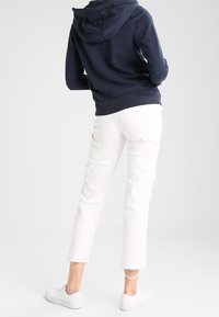 Marineblauwe hoodie met een trekkoord en kangoeroezak, gecombineerd met witte cropped jeans met rafelige zomen en witte sneakers.